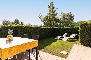 Um pátio externo com mesa e cadeiras, um hedge e uma área gramada.