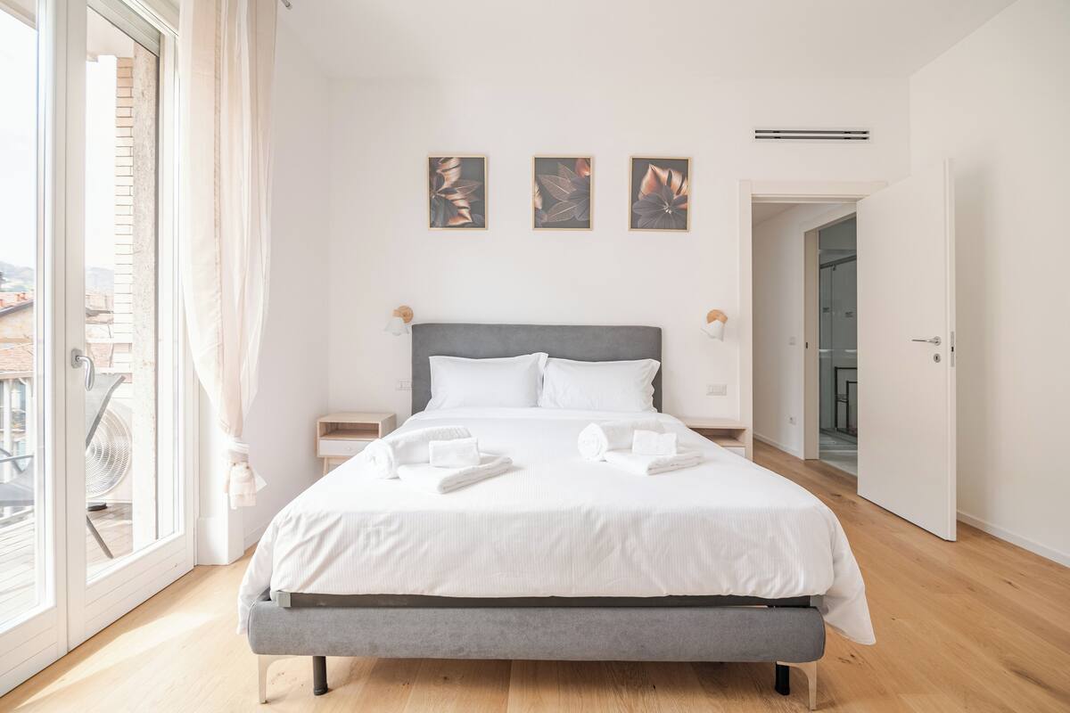 A modern bedroom with a grey bed, white bedding, and three framed floral artworks on the wall.