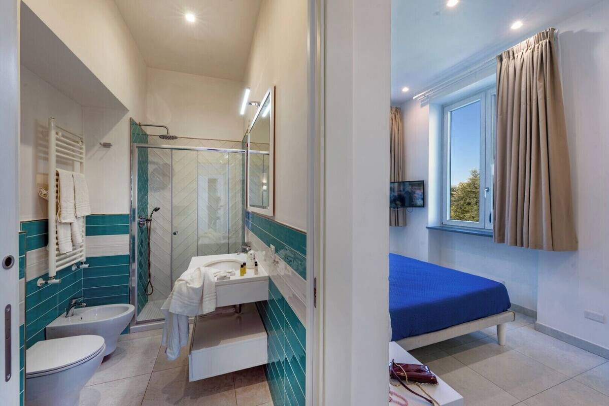 Un baño con ducha, inodoro y lavamanos. Hay una cama con colchón azul y una ventana con cortinas.