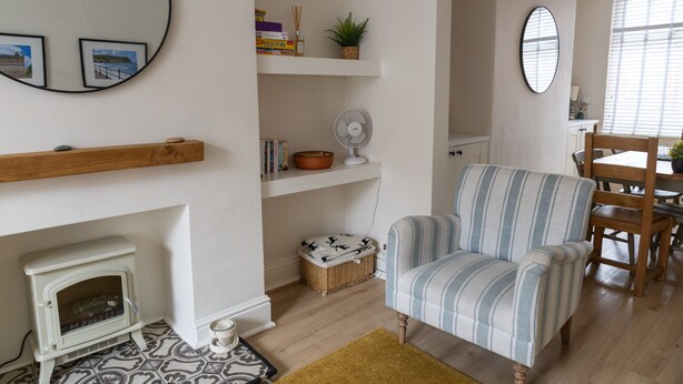 Ein gemütliches Wohnzimmer mit Kamin, einem gestreiften Sessel, einem Esstisch mit Stühlen und einem großen Fenster mit Jalousien.