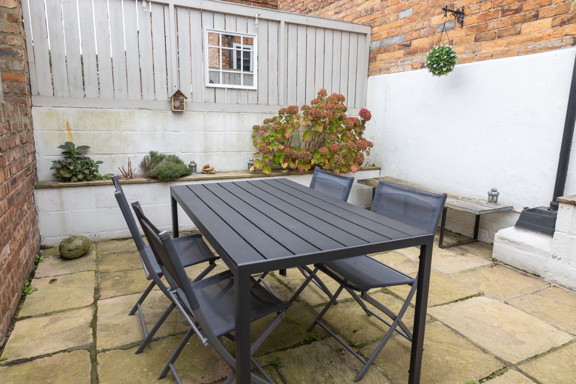 Eine Terrasse mit einem Metalltisch und -stühlen, einer Backsteinmauer, einem weißen Zaun und einem kleinen Garten mit Pflanzen und einem Vogelhaus.