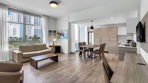 A modern living room with a sofa, coffee table, dining table, and chairs. There is a kitchen area with cabinets and a TV. Large windows provide a view of the cityscape. - Between Lakes and Ocean For 4 (Hollywood)