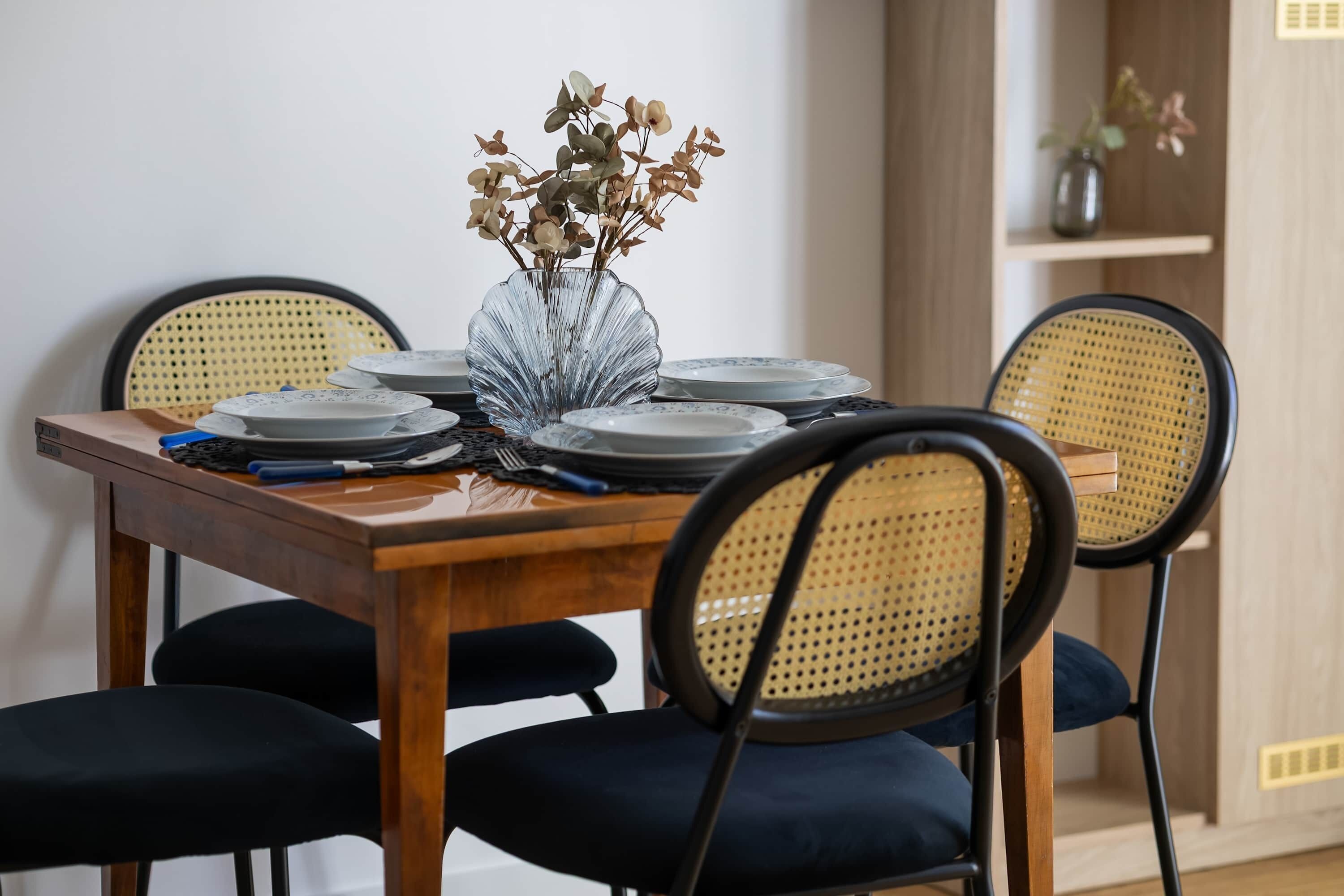 A dining table set with plates, a vase with dried flowers, and black chairs with woven backs.