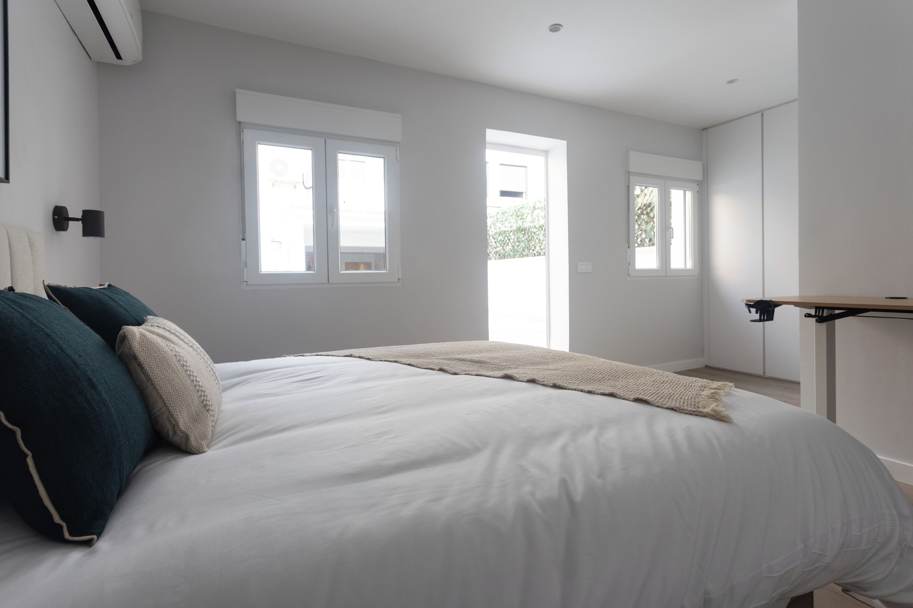 A bedroom with a bed, two windows, a door, and a bedside table.