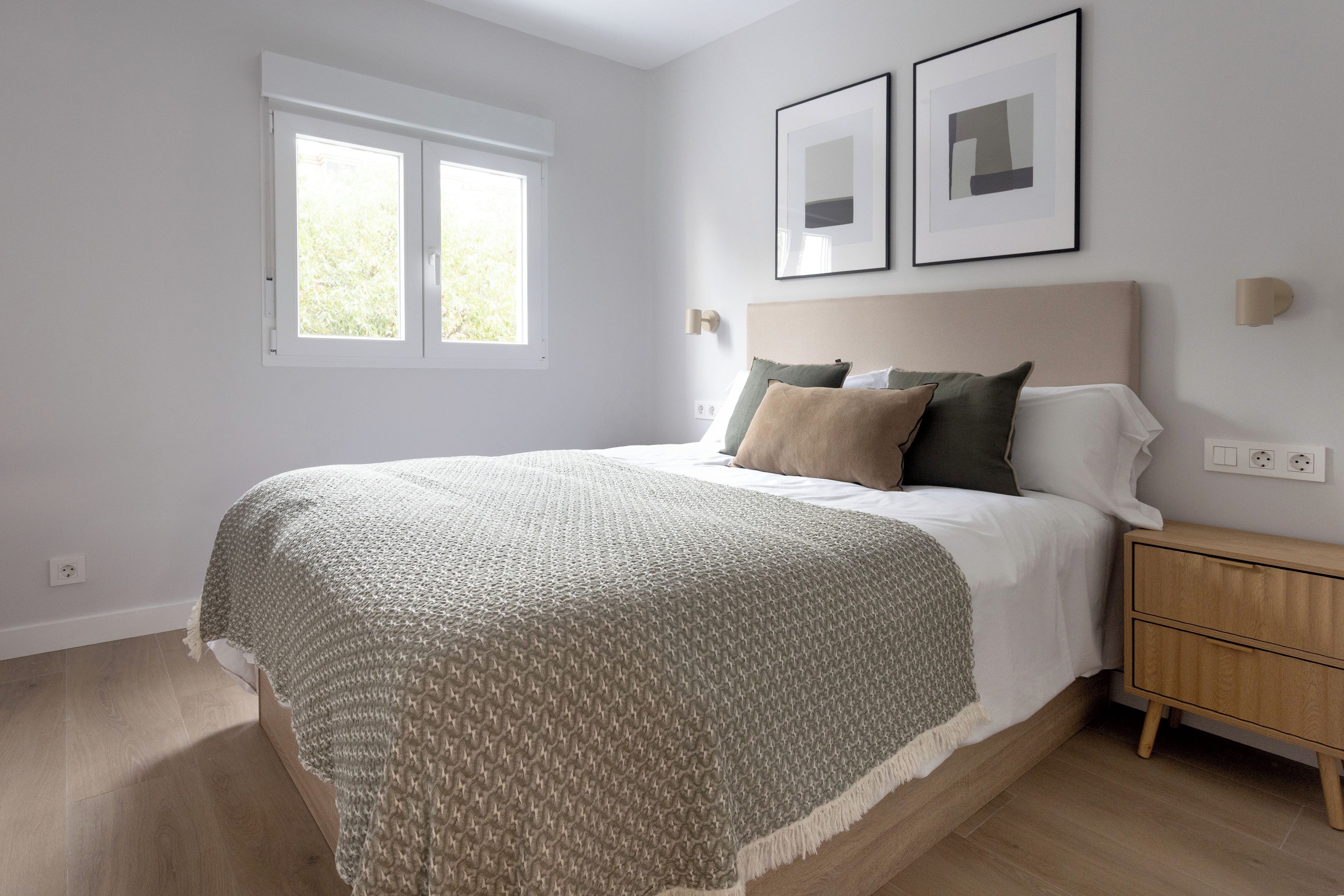 A bedroom with a bed, bedside table, wall-mounted light, and two framed pictures.