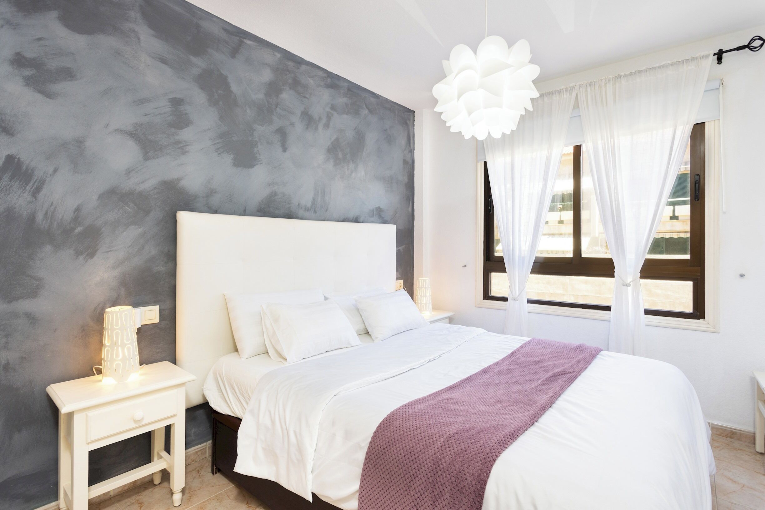 A modern bedroom with a large bed, a bedside table with a lamp, and a view of the outdoors through sheer curtains.