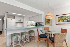A modern kitchen with white cabinets, a microwave, and a sink. There is a dining area with a glass table and wicker chairs. - Seaward B101 (New Smyrna Beach)