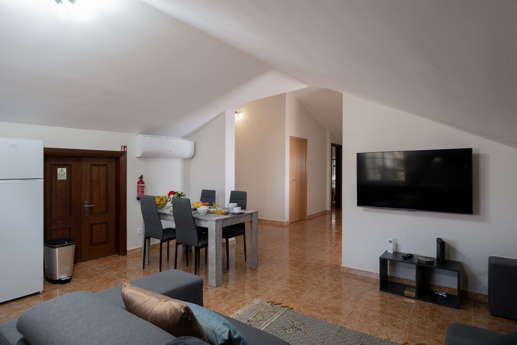 A modern living room with a flat-screen TV, a dining area with chairs and a table, and a kitchen with a refrigerator and a trash bin.