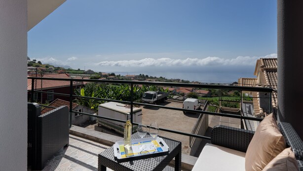 A balcony with wicker furniture, a table with a tray, and a view of houses and a truck in the distance. - 2-bed apt with sea view |Ponta do Sol SeaView (Socorro)