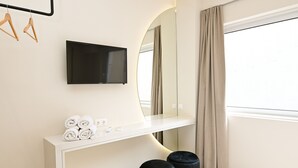 Habitación de hotel moderna con televisión de pantalla plana montada en una consola blanca, un espejo grande y una ventana con cortinas.