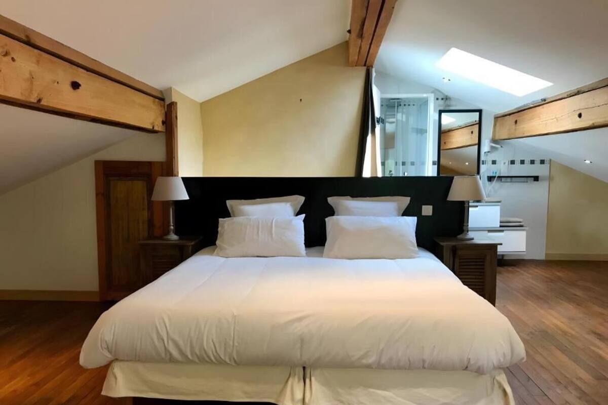 Une chambre spacieuse avec un grand lit, des poutres apparentes, une lucarne et une salle de bain moderne.