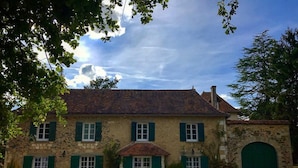 Un bâtiment en pierre doté d’une entrée centrale, de nombreuses fenêtres avec volets et d’une grande porte en arc de cercle.
