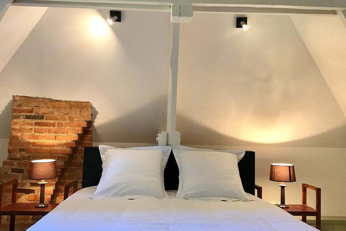 Une chambre avec un plafond haut, des poutres apparentes, un mur en briques, un lit recouvert de draps blancs, deux tables de chevet avec des lampes et une petite fenêtre.