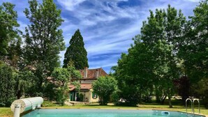 Une piscine rectangulaire avec une terrasse en bois, entourée d’arbres et, au fond, une maison avec un toit rouge.
