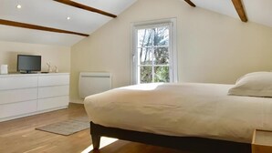 Een slaapkamer met een schuine kap, een bed, een dressoir, een televisie en een raam met uitzicht op bomen.