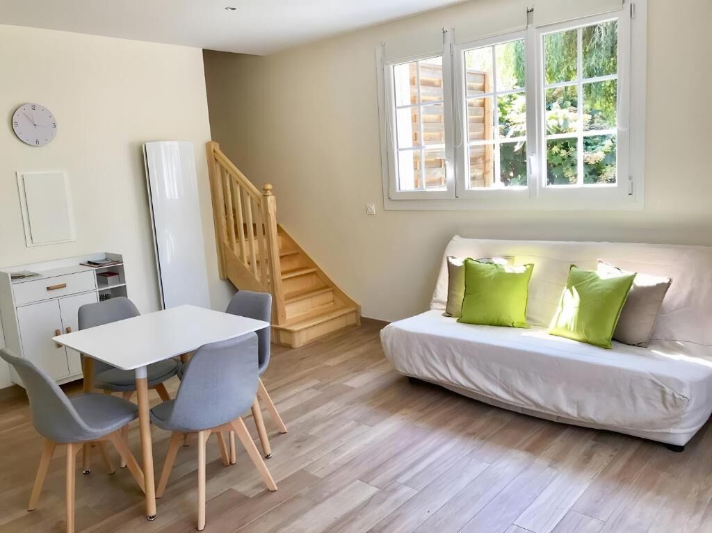 Um quarto com um sofá branco, uma mesa de jantar com cadeiras, uma escada e uma janela com vista para o verde.