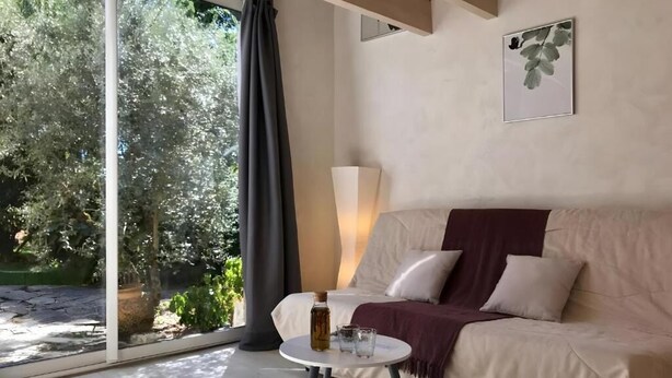Une pièce avec un canapé beige, une petite table ronde sur laquelle se trouvent une bouteille et des verres, un lampadaire et une vue sur la verdure par une grande fenêtre.