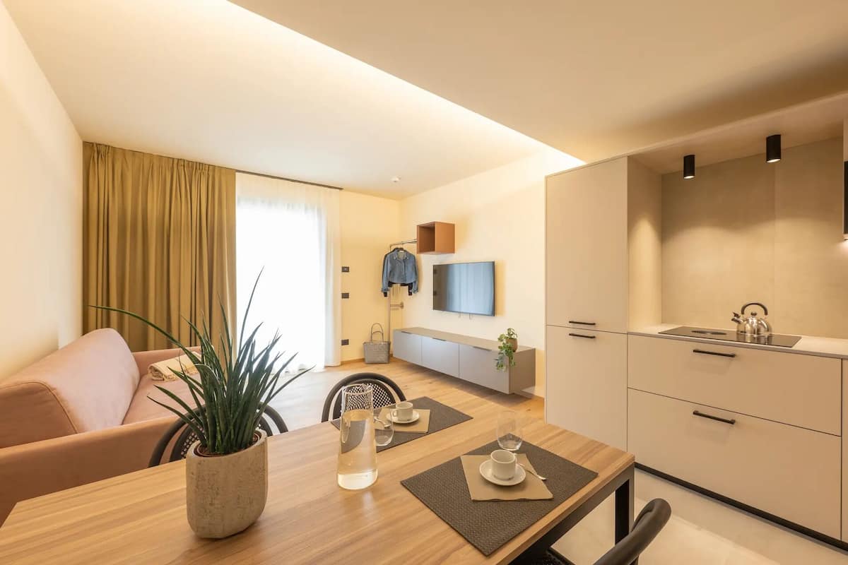 Un salon moderne comprenant un canapé, une table à manger avec des sets de table, une table basse avec une plante, et un coin cuisine avec des placards et un évier.