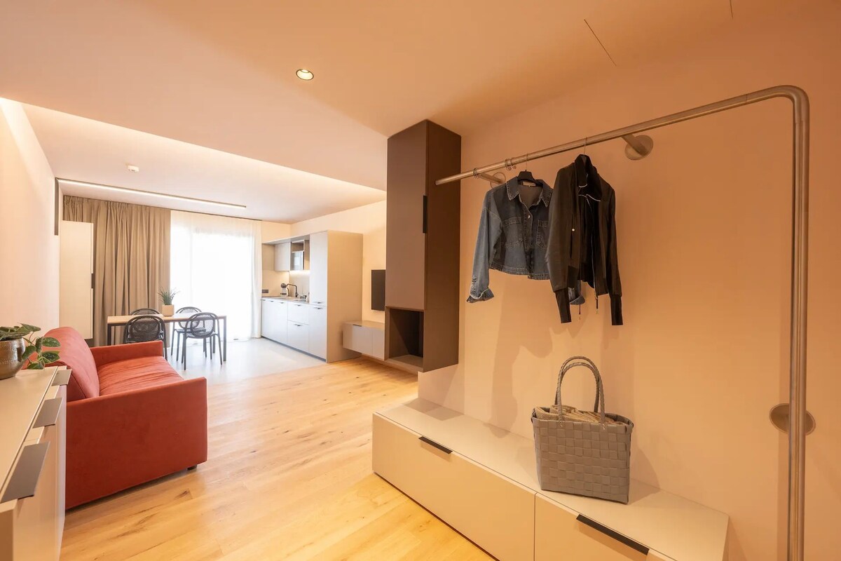 A modern hotel room with a wooden floor, a red sofa, a dining area, a kitchen, and a clothing rack with jackets and a handbag.