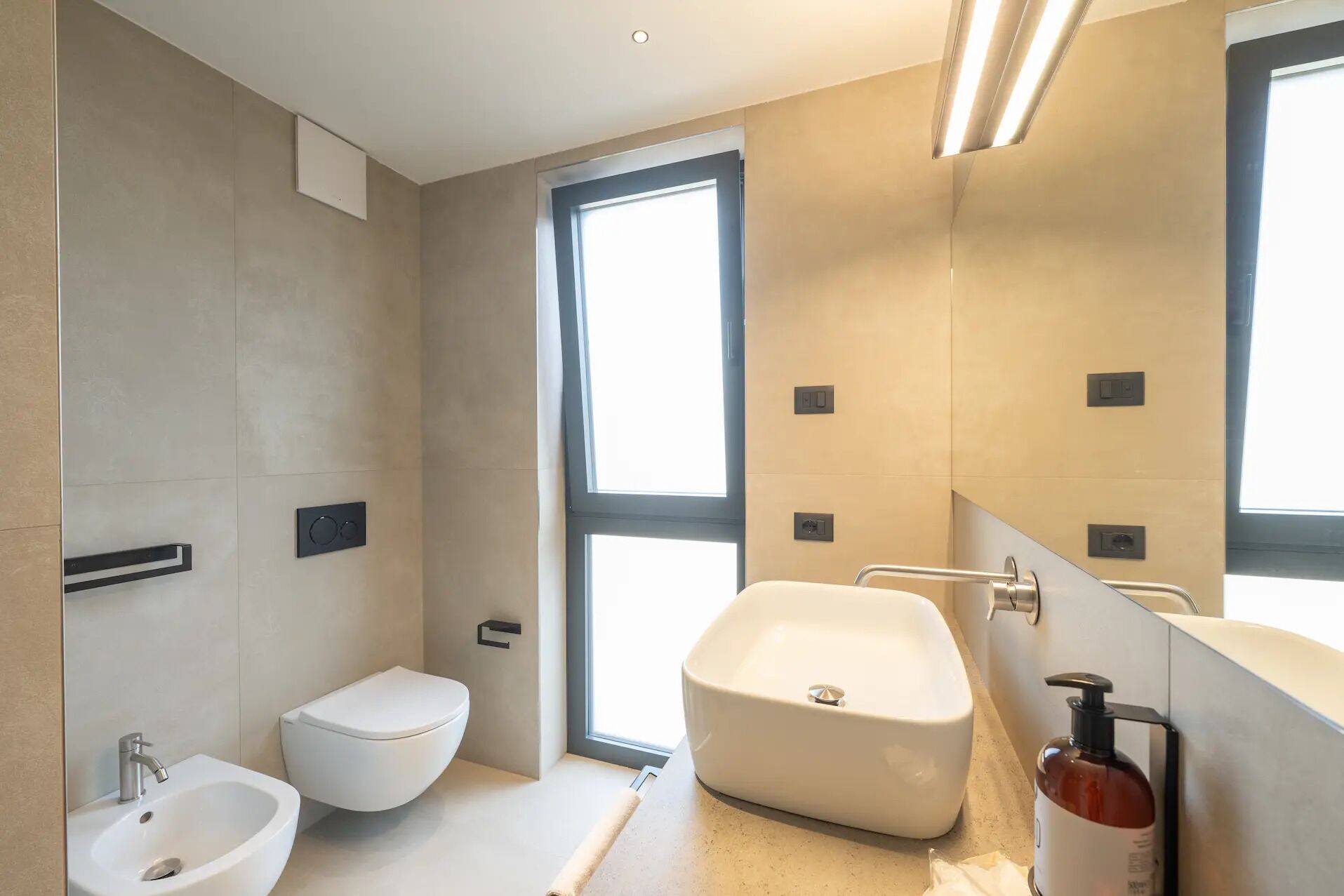 A modern bathroom with a toilet, bidet, and bathtub, large windows, and a mirror.