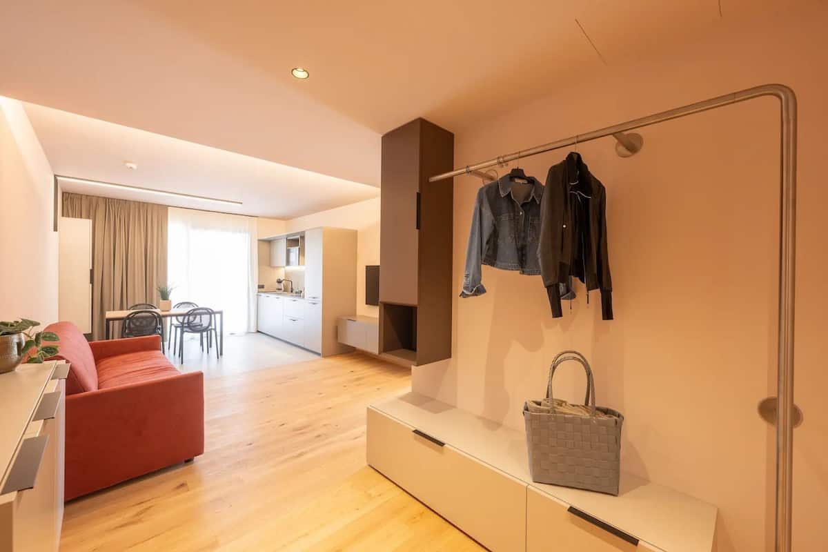 Une chambre d’hôtel moderne avec un plancher en bois, un canapé rouge, un coin repas, une cuisine et un portant à vêtements avec des vestes et un sac à main.
