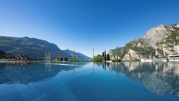 Una piscina azul cristalina con montañas al fondo, una terraza de madera con sillones de descanso y un acantilado rocoso.