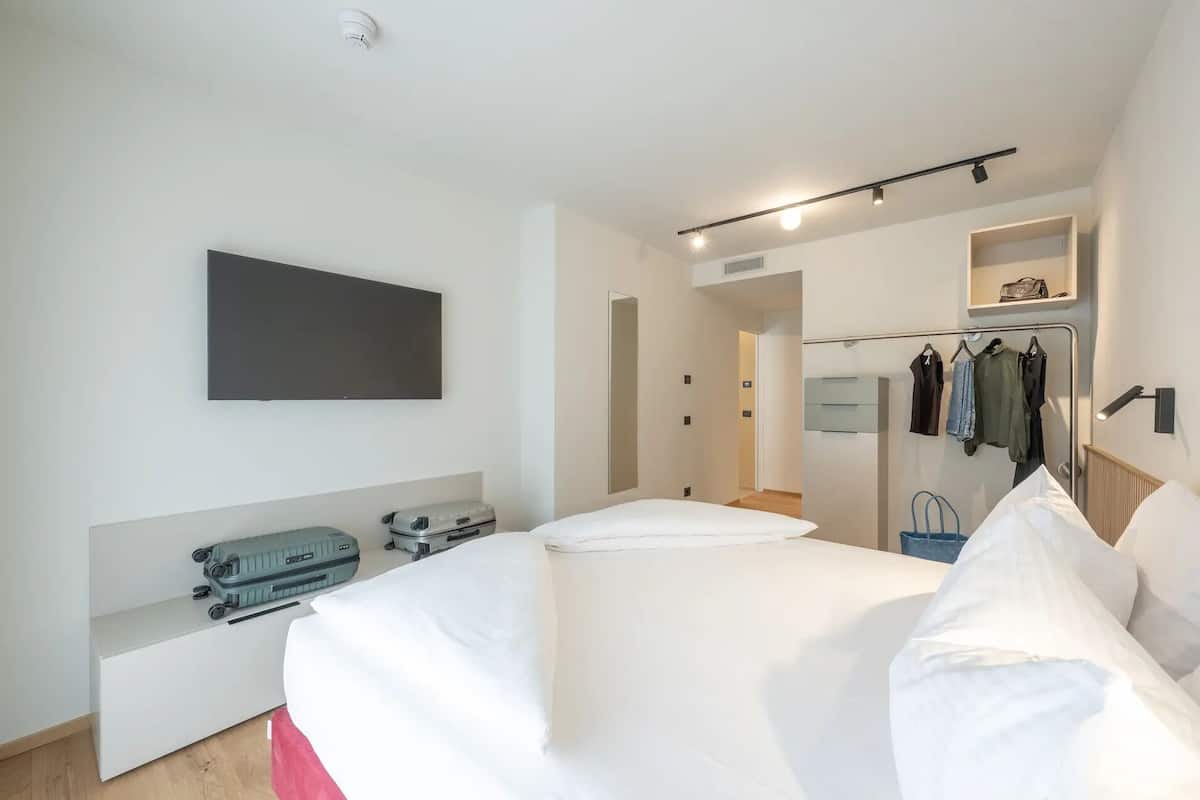 Une chambre d’hôtel avec un lit, un téléviseur à écran plat fixé au mur, un porte-manteau avec des vêtements et une valise posée sur le sol.
