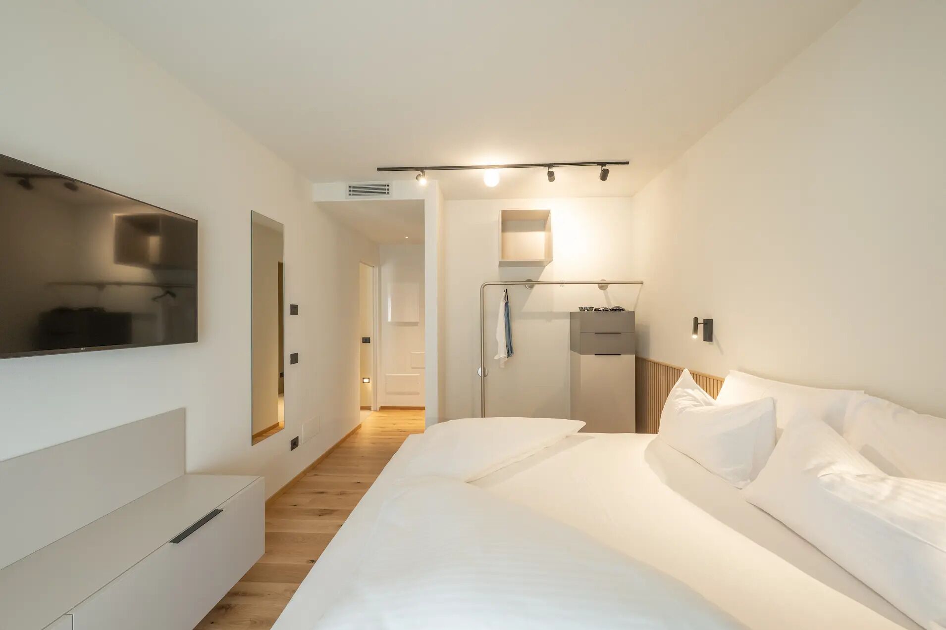Una habitación de hotel moderna con una cama grande, un televisor de pantalla plana montado en la pared, piso de madera y un baño con ducha y toallero.