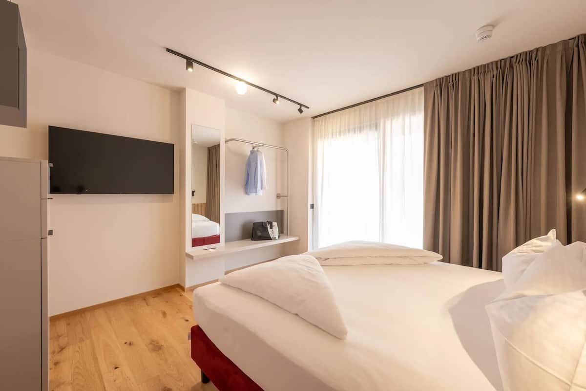 Une chambre d’hôtel avec un lit, un téléviseur fixé au mur, un miroir, une étagère avec une serviette et une tasse, et une fenêtre avec des rideaux.