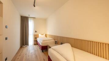 A hotel room with a wooden floor, a bed with white bedding, a bedside table, and a window with curtains.