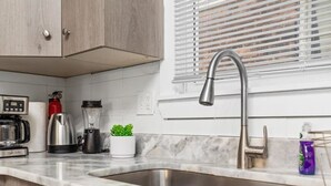 A kitchen with a stainless steel sink, wooden cabinets, a coffee maker, a kettle, a blender, and a window with blinds. - The Sheridan Family Compound 4BR + 2.5BA (Detroit)