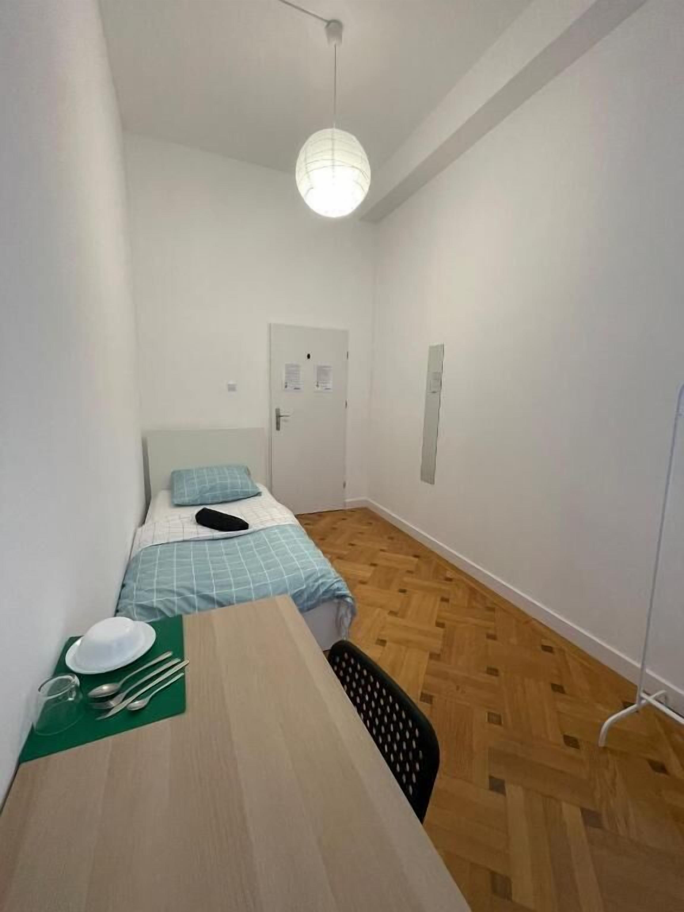 A small, clean room with a bed, a desk, and a chair. There is a door and a light fixture on the ceiling.