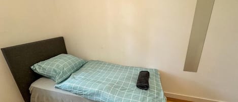 Ein kleines Zimmer mit einem Einzelbett, einem dunklen Kopfteil, einer hellen Matratze und einer blau-weiß karierten Tagesdecke.