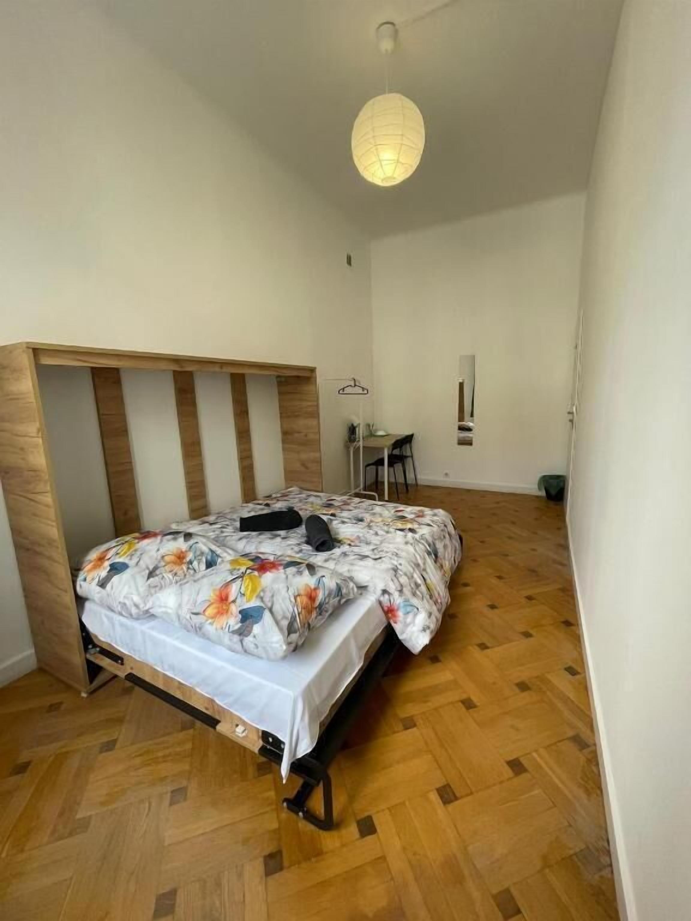 A bedroom with a wooden headboard, a bed with a floral duvet, a hanging light fixture, and a small desk with a chair.