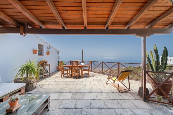 A covered patio with a table and chairs, potted plants, and a view of the sea.