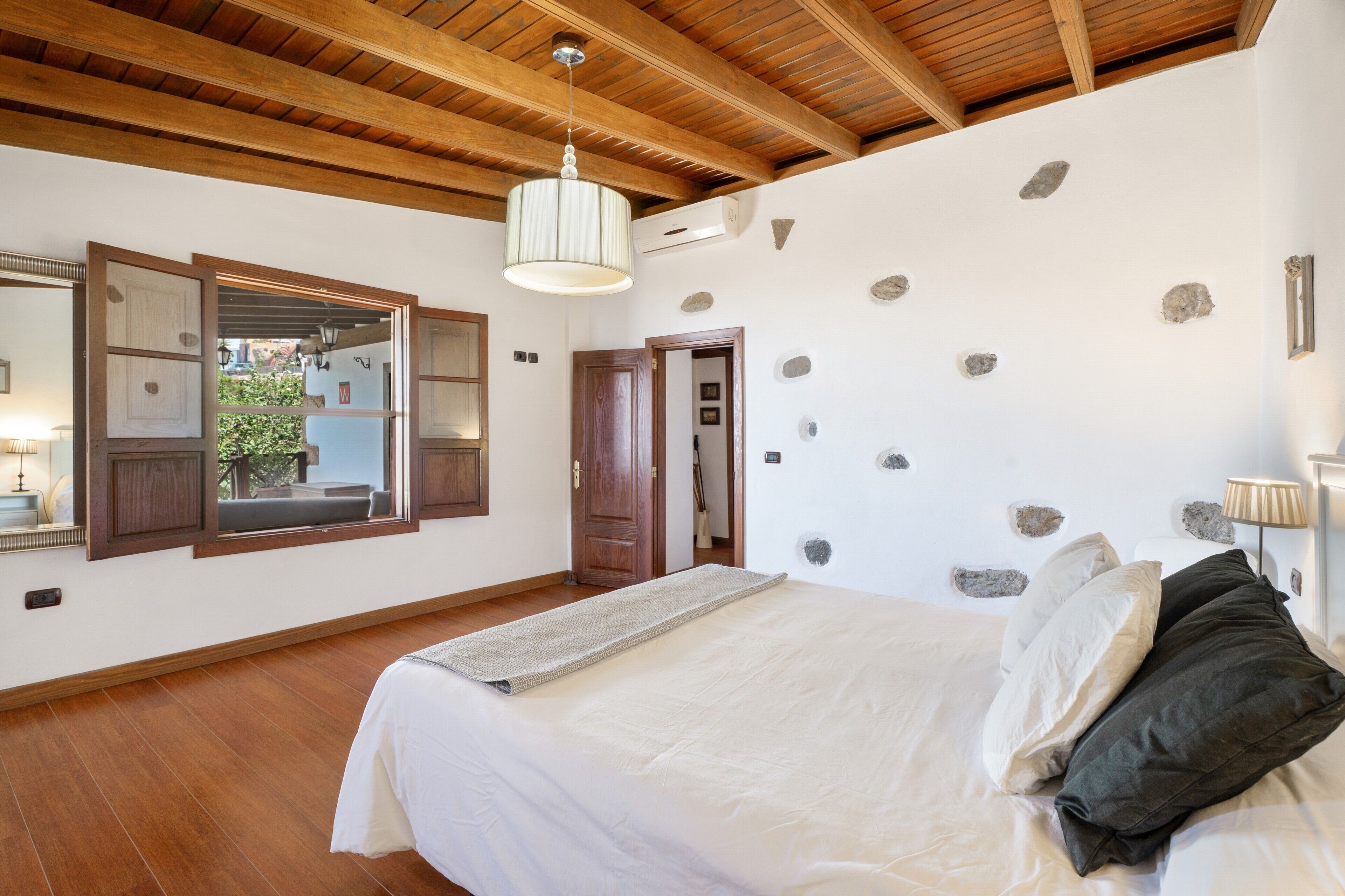 A bedroom with a wooden ceiling, a bed with white bedding, a mirror, and a stone wall.