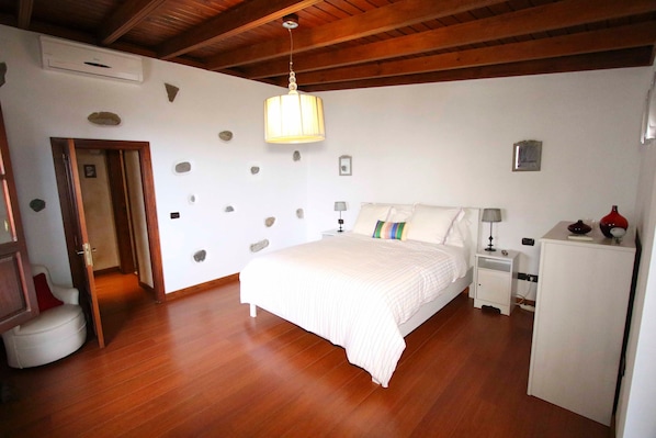 A bedroom with a bed, nightstands, a dresser, a hanging light fixture, and a door leading to another room.