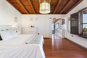 A bedroom with a large bed, a bedside table, a lamp, and a view into the adjacent kitchen.