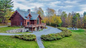 A log cabin with a stone foundation, a wooden deck, and a paved driveway leading to a circular seating area. - Spruce Moose Lodge, Custom Log Cabin with Hot Tub (Hartland)
