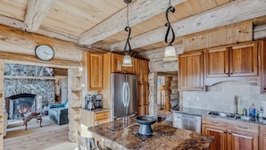 A rustic kitchen with wooden cabinets, a granite countertop, a stainless steel refrigerator, and a stone fireplace. - Spruce Moose Lodge, Custom Log Cabin with Hot Tub (Hartland)