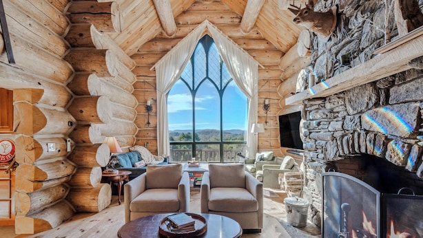 A log cabin interior with a stone fireplace, two armchairs, a coffee table, and a large window with a view of the outdoors. - Spruce Moose Lodge, Custom Log Cabin with Hot Tub (Hartland)