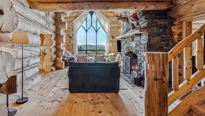 A log cabin interior with a stone fireplace, wooden ceiling, and a view through a large window. - Spruce Moose Lodge, Custom Log Cabin with Hot Tub (Hartland)