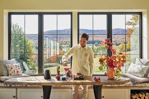 Une personne verse une boisson à une table, avec vue sur un paysage boisé à travers de grandes fenêtres.