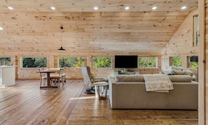 A spacious cabin interior with a dining area, sofa, and large windows offering views of greenery.