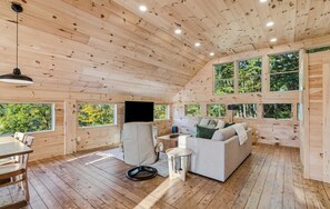A wooden cabin interior with a sofa, a dining table, and multiple windows offering views of the forest.