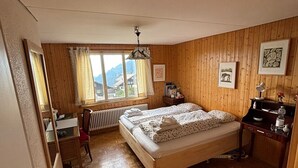 Ein Schlafzimmer mit Holzboden, einem Bett mit gemustertem Überwurf, einem Schreibtisch mit Stuhl, einem Heizkörper und einem Fenster mit Vorhängen.