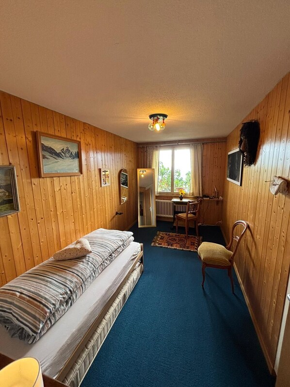 A room with wooden walls, a bed, a desk, a chair, and a window.