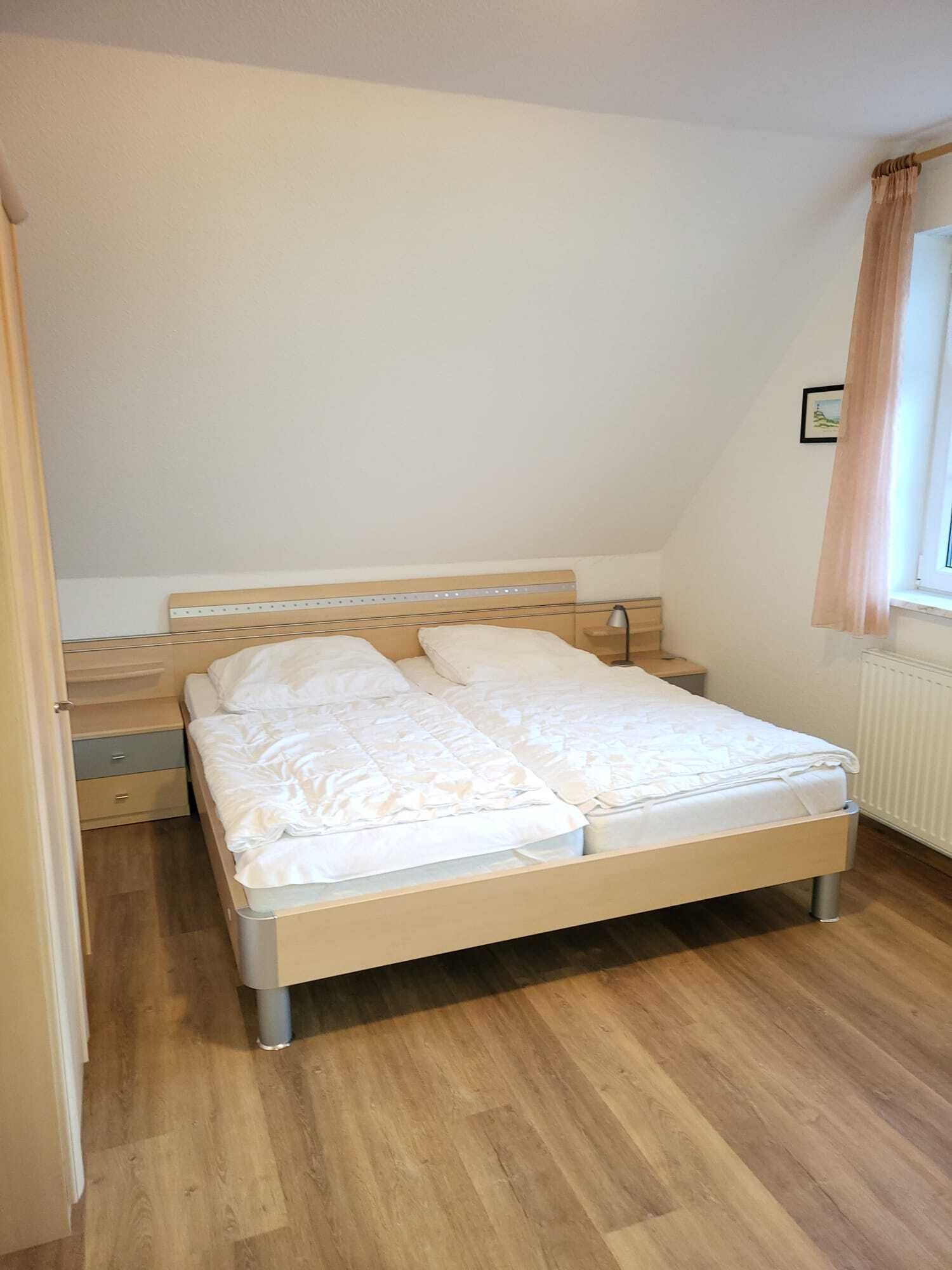 Ein kleines Zimmer mit einem Einzelbett, einem Holzkopfstück, einem Nachttisch und einem Fenster mit Vorhängen.