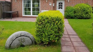 A brick house with a red-tiled roof, a white door, and a large window. There is a stone marker with the number 13, a paved pathway, and well-maintained grass. - House Kiki in Dornumersiel (Dornum)