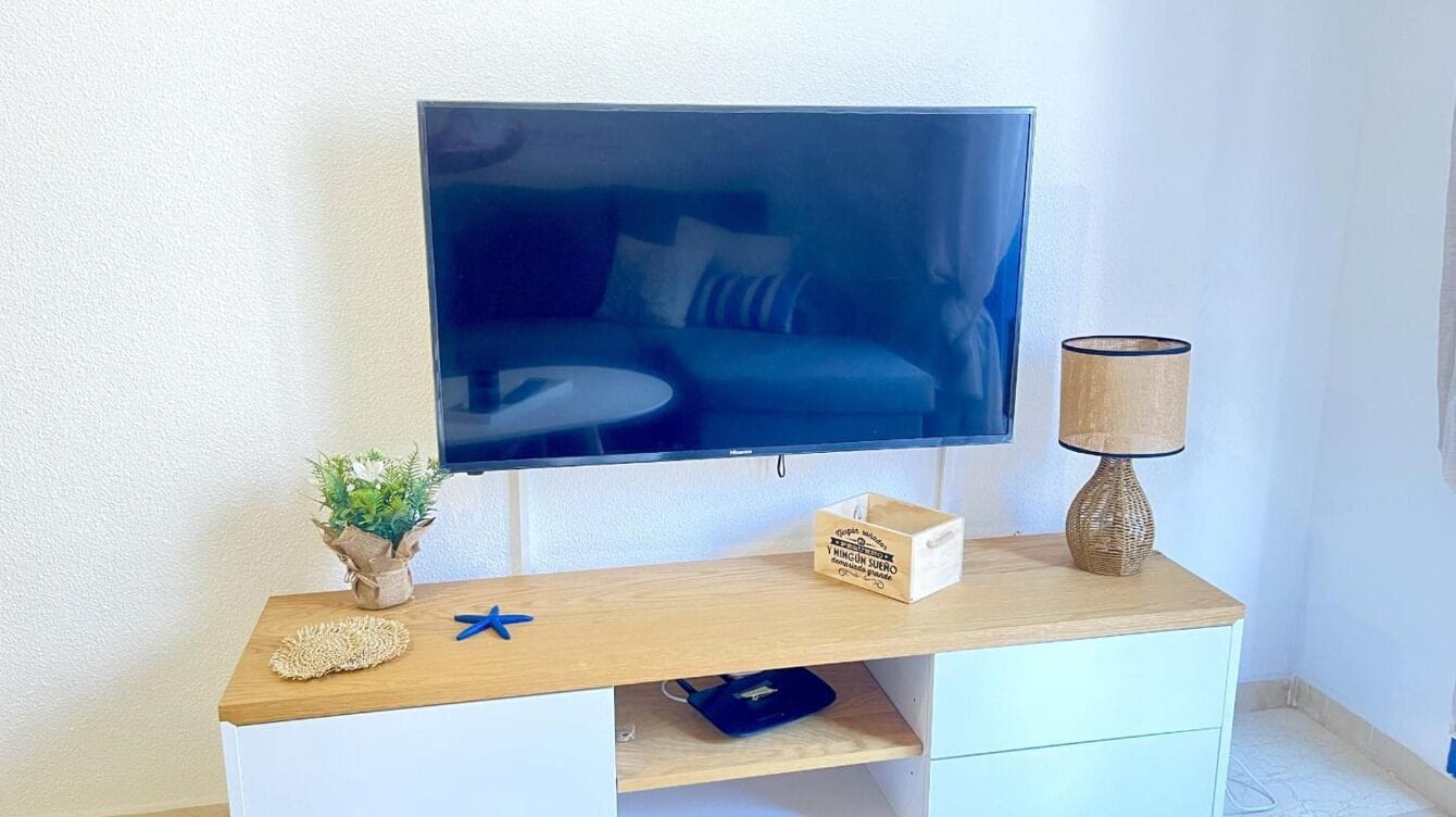 Un téléviseur à écran plat fixé au mur, au-dessus d’une console en bois sur laquelle se trouvent une lampe, une petite plante et un objet bleu en forme d’étoile.